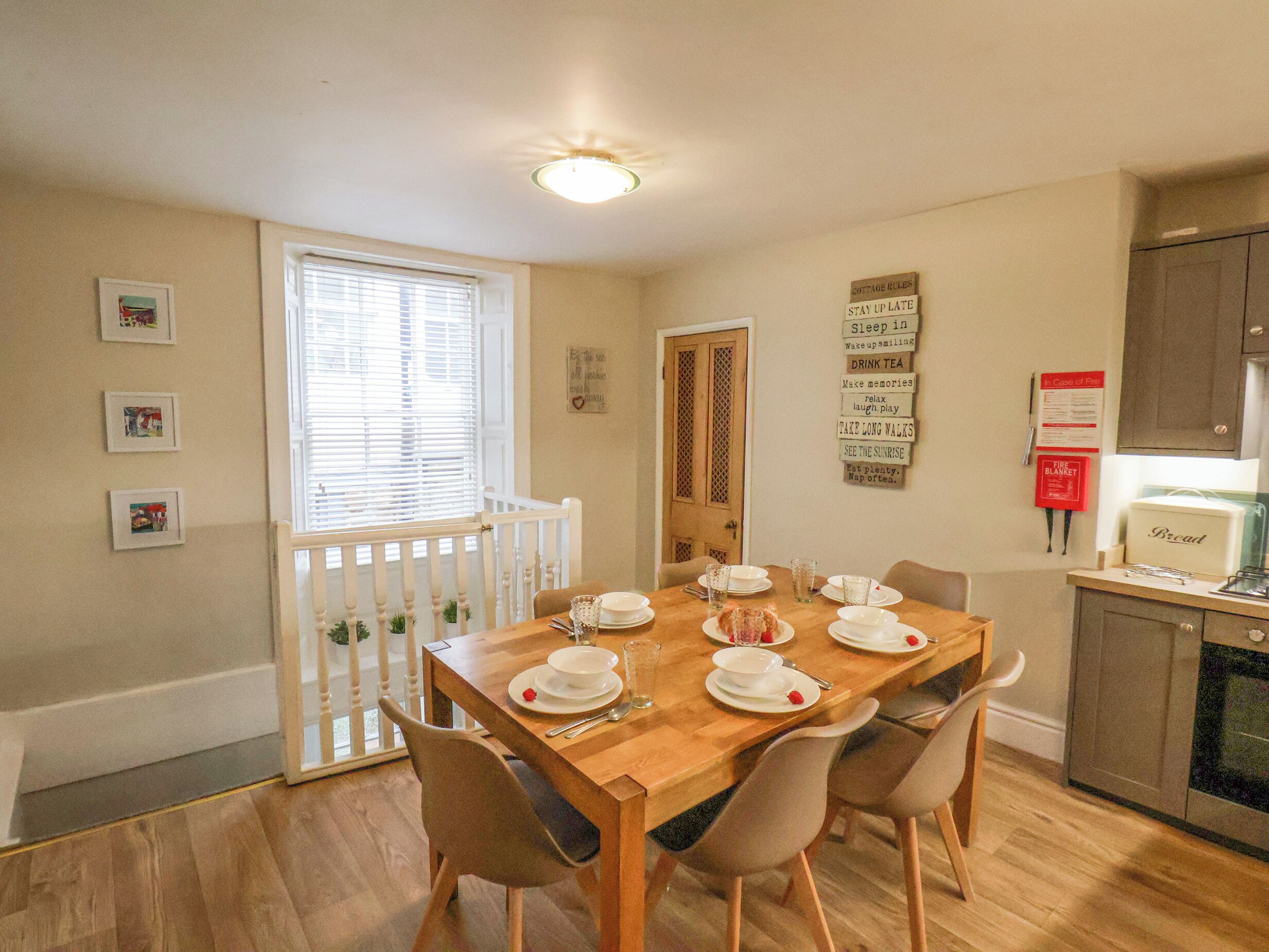 A dining area with a wooden table set for four, surrounded by chairs. There are framed pictures on the wall, a window with blinds, and a door with a decorative panel.
