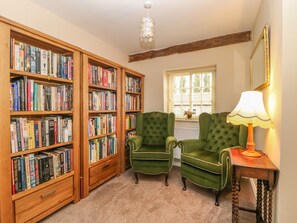 A cozy reading room with wooden bookshelves filled with books, two green armchairs, a table with a lamp, and a window with blinds. - Copmanthorpe Hall (York)