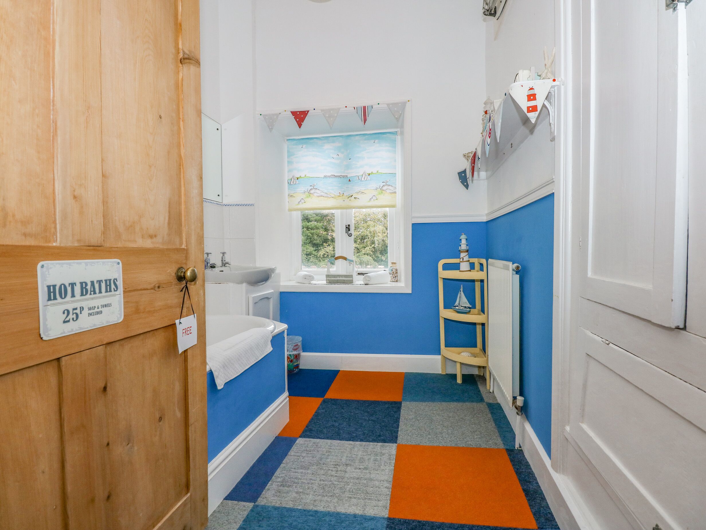Ein Badezimmer mit blauer und weißer Farbgestaltung, ein Fenster mit Blick auf Grünflächen, ein Waschbecken, eine Toilette und ein Schild, das ankündigt, dass heiße Bäder für 25 Pence verfügbar sind.