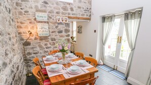 A dining area with a stone wall, a wooden table set for four, and a view of the outside through a door with curtains. - Castle School House (Kidwelly)