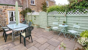 A stone house with a patio area featuring a table and chairs, a green fence, and potted plants. - Castle School House (Kidwelly)