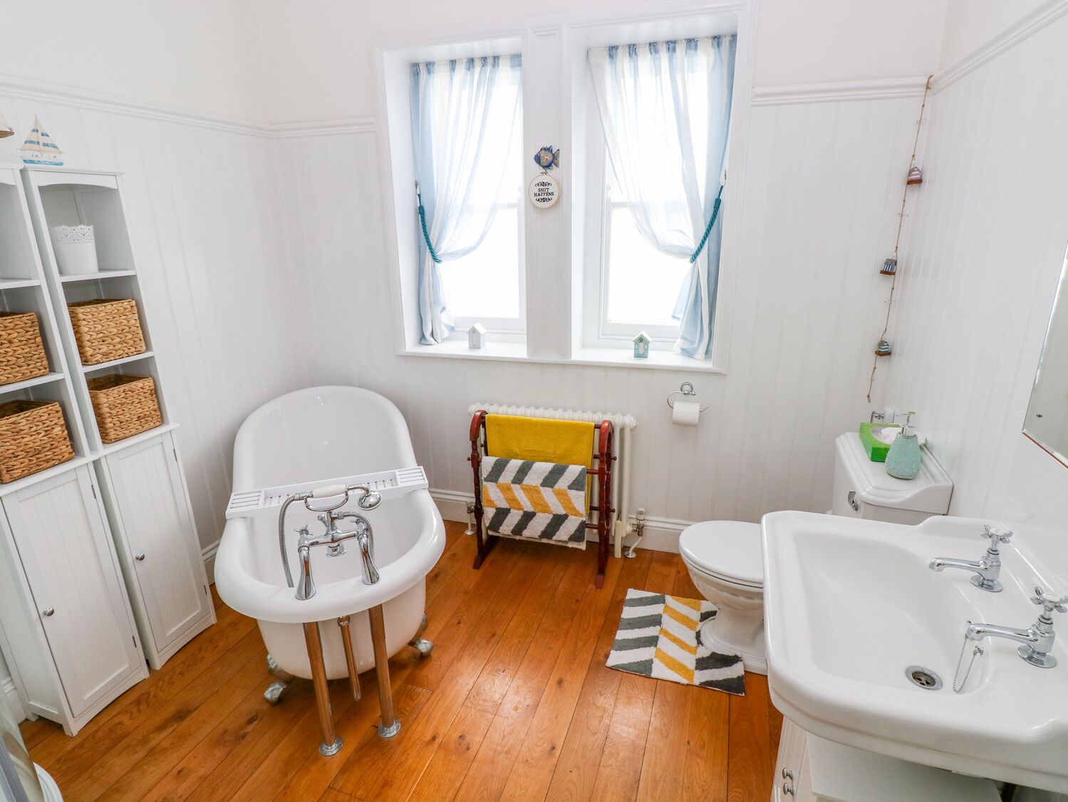 Een badkamer met een witte badkuip, een witte wastafel en een toilet. Er is een raam met gordijnen, een plank met manden en een radiator.