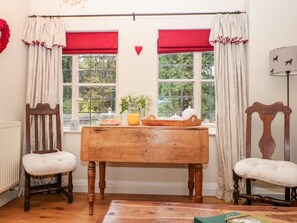 A cozy room with a wooden table, two chairs, a radiator, and two windows with red curtains. - October Cottage (Stroud)