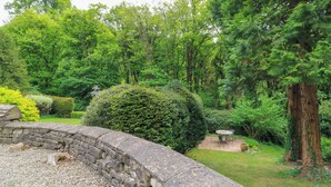 A stone wall with a circular stone table and chairs, surrounded by lush greenery and trees. - October Cottage (Stroud)