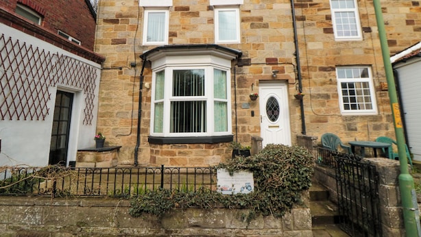 A two-story stone house with a bay window, a white door, and a small garden area with a fence.