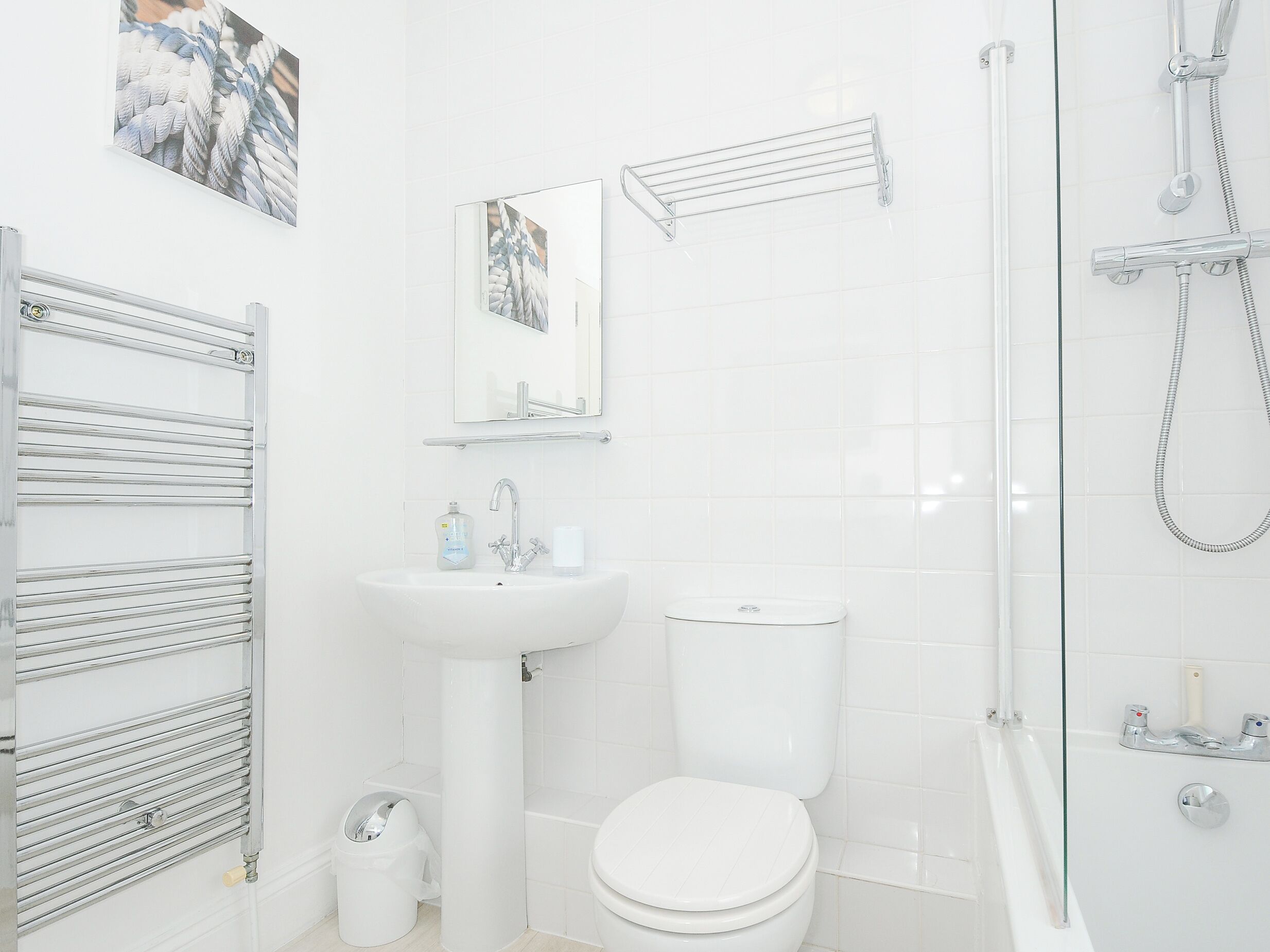 Ein modernes Badezimmer mit Waschbecken, Toilette und Dusche in Weiß. Handtuchhalter, Spiegel und eine Duschkabine aus Glas sind vorhanden.