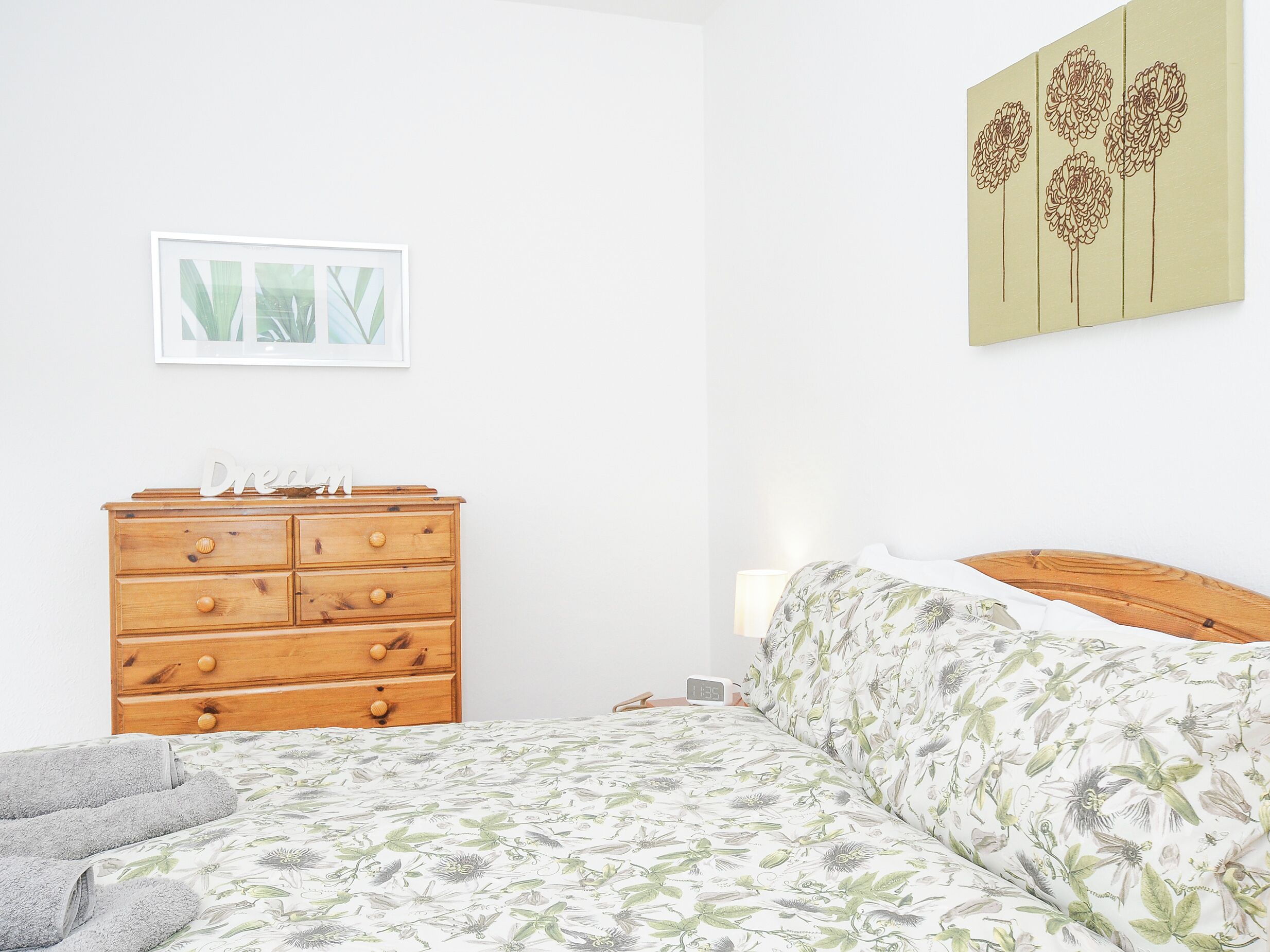 Un dormitorio con un aparador de madera, una cama con ropa de cama floral, una mesita de noche, una lámpara y un cuadro en la pared.