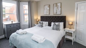 A neatly made bed with white linens and two pillows, a bedside table with a lamp, a window with curtains, and a view of buildings outside. - Tennyson House (Scarborough)