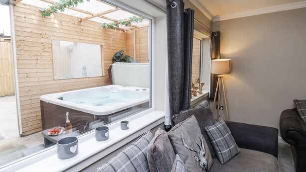 A cozy room with a large window, a sofa with grey cushions, a small table with a bowl of fruit and mugs, and a view of an outdoor wooden deck and hot tub. - Tennyson House (Scarborough)
