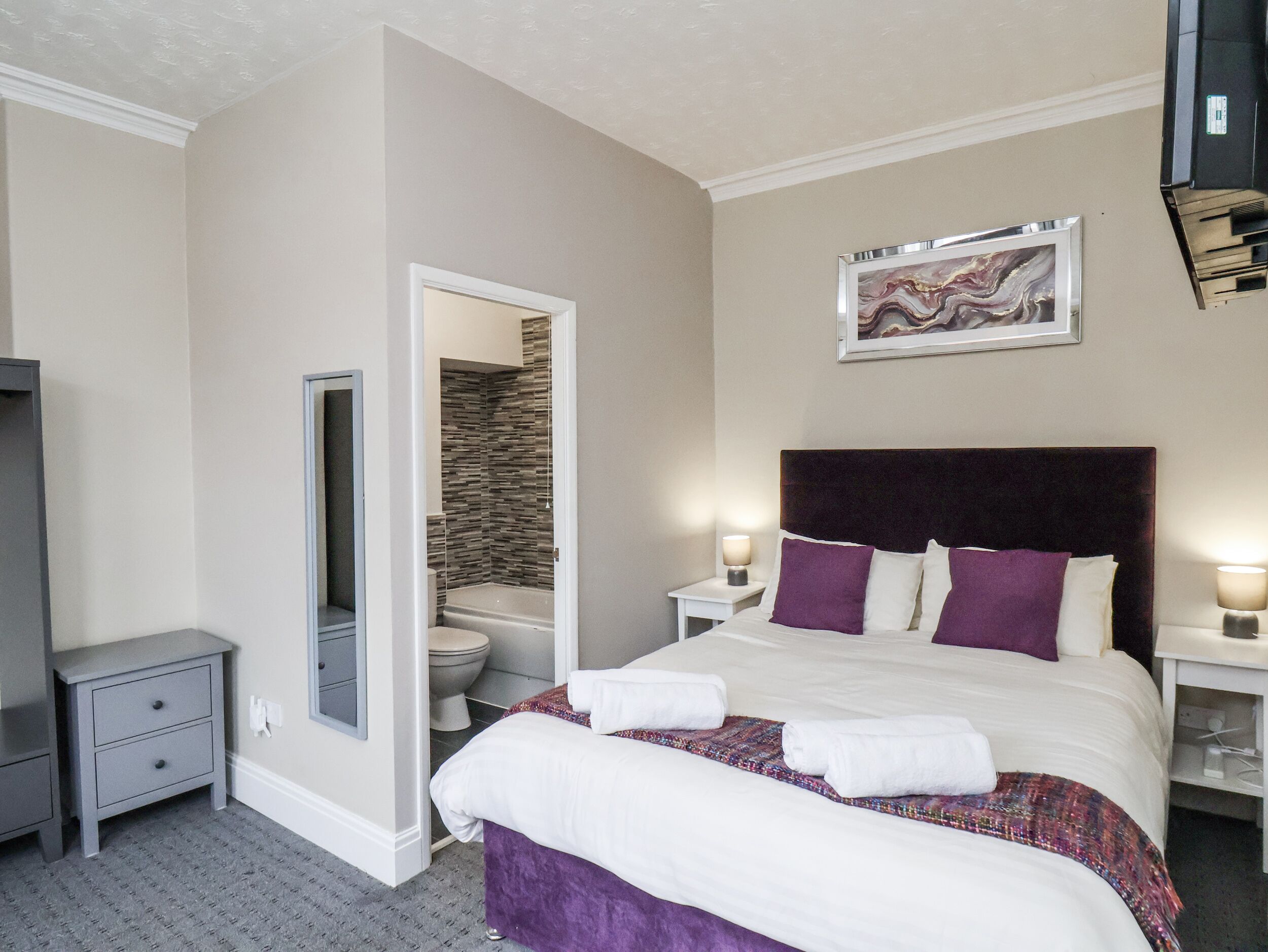 Quarto de hotel com cama, mesa de cabeceira, cômoda, banheiro com vaso sanitário e chuveiro, e uma obra de arte emoldurada na parede.