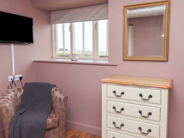 A room with a television mounted on the wall, a window with a view of the outdoors, a mirror with a gold frame, and a white dresser with black handles. - The Cottage (Thirsk)