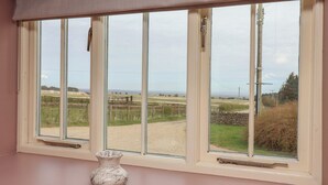A window with a view of a rural landscape, a white vase on the windowsill, and a beige curtain. - The Cottage (Thirsk)