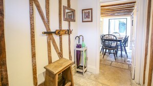 Un intérieur rustique avec des poutres apparentes, un tabouret en bois, une petite table et un coin repas avec des chaises et une table visibles à travers une porte ouverte.