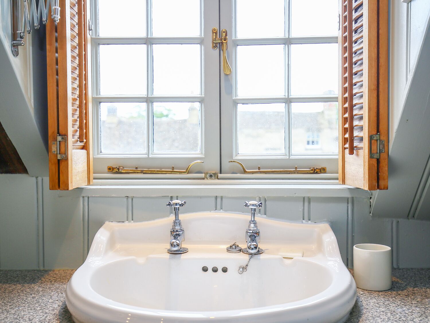 A bathroom with a large white sink, two faucets, and a window with wooden shutters.