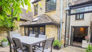 Un bâtiment en pierre avec un toit en tuiles, une porte d’entrée en verre et un patio aménagé avec une table et des chaises.