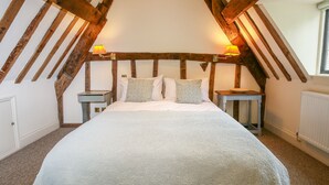 Une chambre avec un plafond en pente, des poutres apparentes, un lit avec une literie blanche, deux tables de chevet et deux lampes fixées au mur.