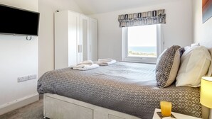 Un dormitorio con cama, armario, televisor, ventana con vistas y mesita de noche con lámpara y vaso de jugo de naranja.