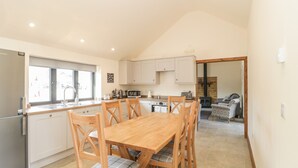 A modern kitchen with a dining area, featuring a wooden table and chairs, a built-in oven, and a large window providing natural light. - The Milking Barn (Yeovil)