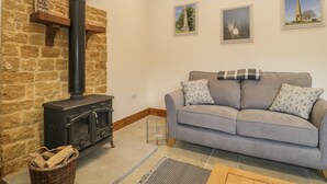 A cozy living room with a stone fireplace, a grey sofa with patterned cushions, and two framed pictures on the wall. - The Milking Barn (Yeovil)