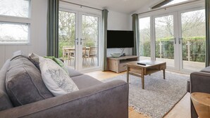 Una sala moderna con un sofá gris, una mesa de centro y un televisor sobre un mueble de madera. Hay una puerta corrediza de vidrio que da a un balcón con una mesa y sillas para comer.