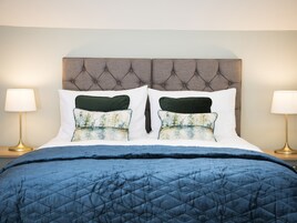 Una cama bien hecha con un edredón azul, dos almohadas blancas y dos almohadas decorativas verdes con estampado floral.