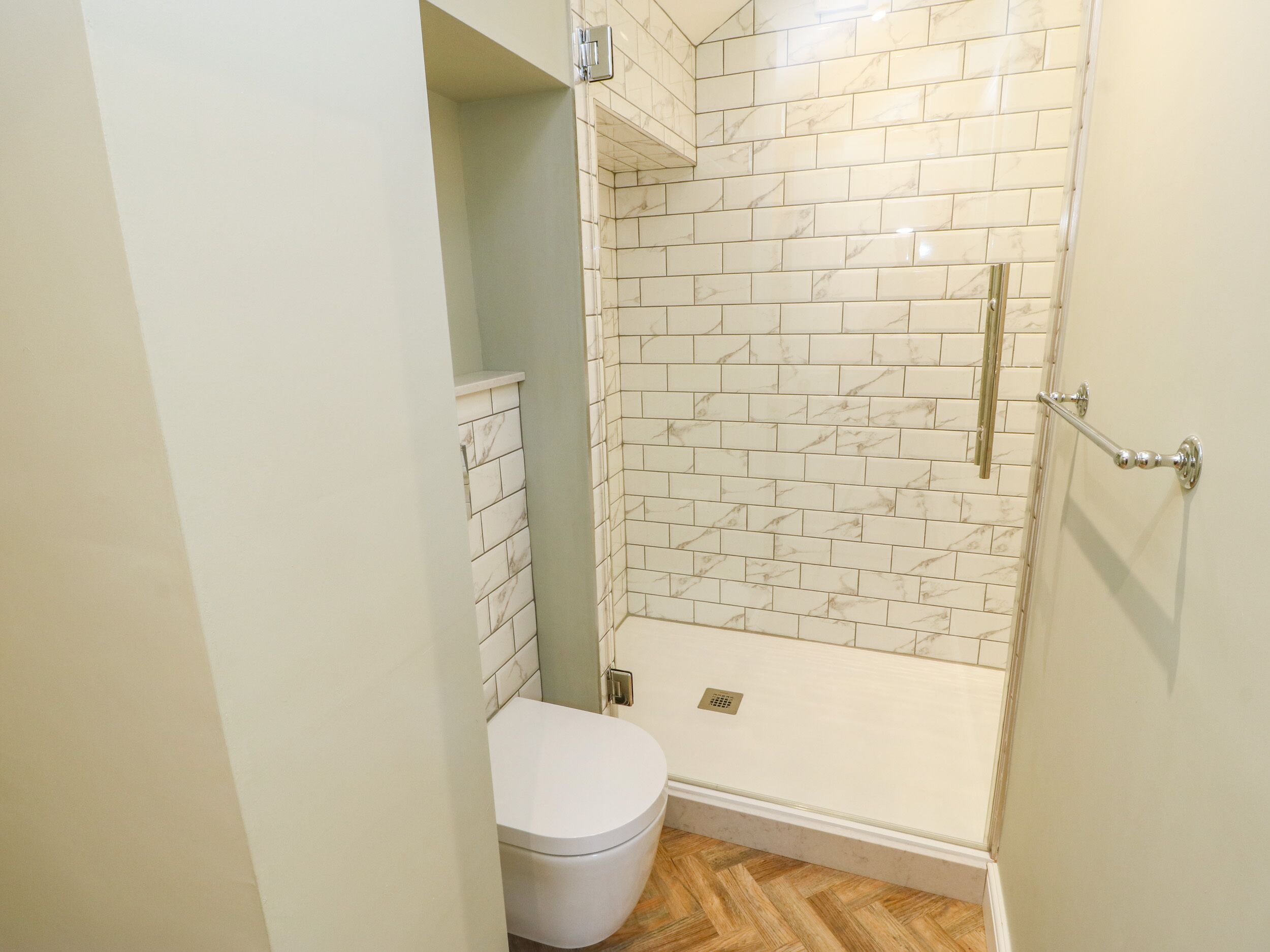 A bathroom with a white toilet, a shower with white tiles, and a wooden floor.