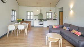 A modern living room with a grey sofa, a wooden dining table with chairs, a small round table, and a kitchen in the background. - Foxglove (Shepton Mallet)