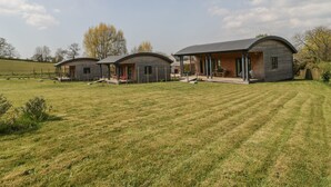 A row of three wooden cabins with curved roofs, each featuring a porch and a small deck. - Foxglove (Shepton Mallet)