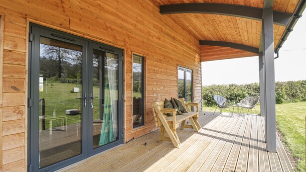 A wooden cabin with large sliding glass doors, a deck with outdoor seating, and a covered patio area. - Foxglove (Shepton Mallet)