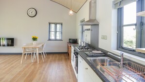 A modern kitchen with wooden flooring, a dining area with a table and chairs, a clock on the wall, and a window with blinds. - Foxglove (Shepton Mallet)