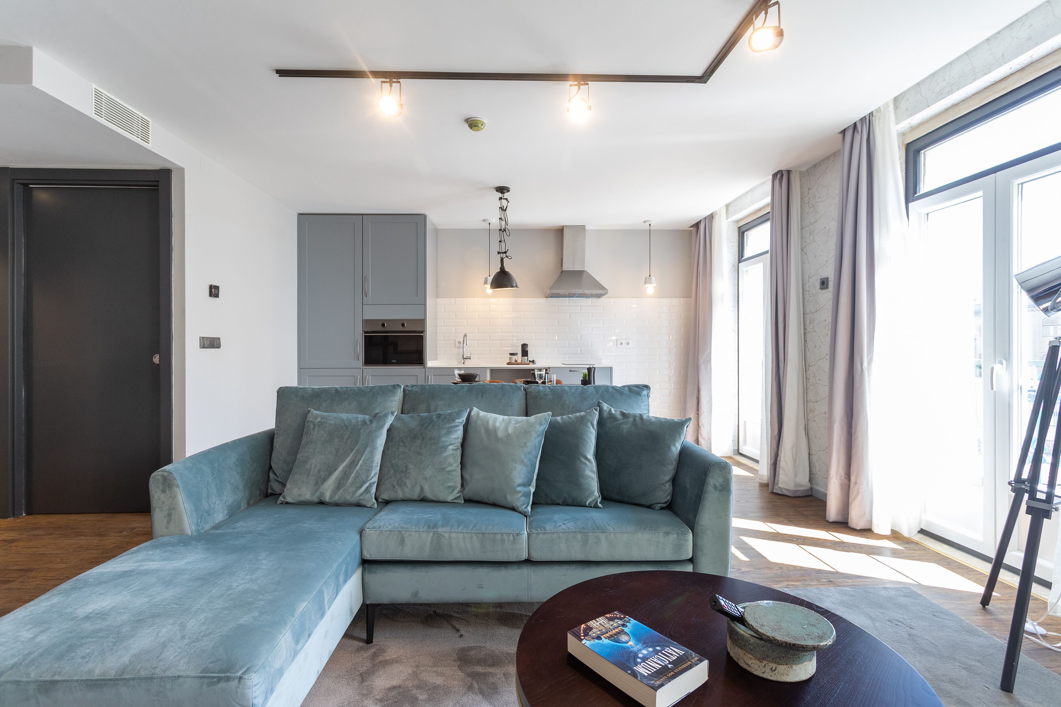 A modern living room with a light blue sectional sofa, a round coffee table with a book and a decorative bowl, and a kitchen in the background with grey cabinets and pendant lighting.