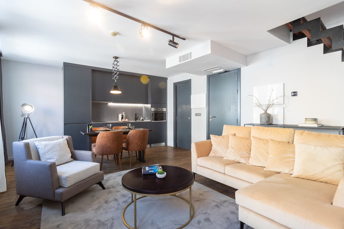 Uma sala de estar moderna com sofá, poltrona e mesa de centro. Há também um espaço de jantar com cadeiras e mesa, e uma cozinha ao fundo.