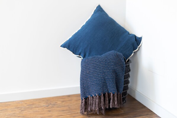 A corner of a room with a wooden floor, a white wall, and a blue textured blanket draped over a stack of dark-colored pillows.