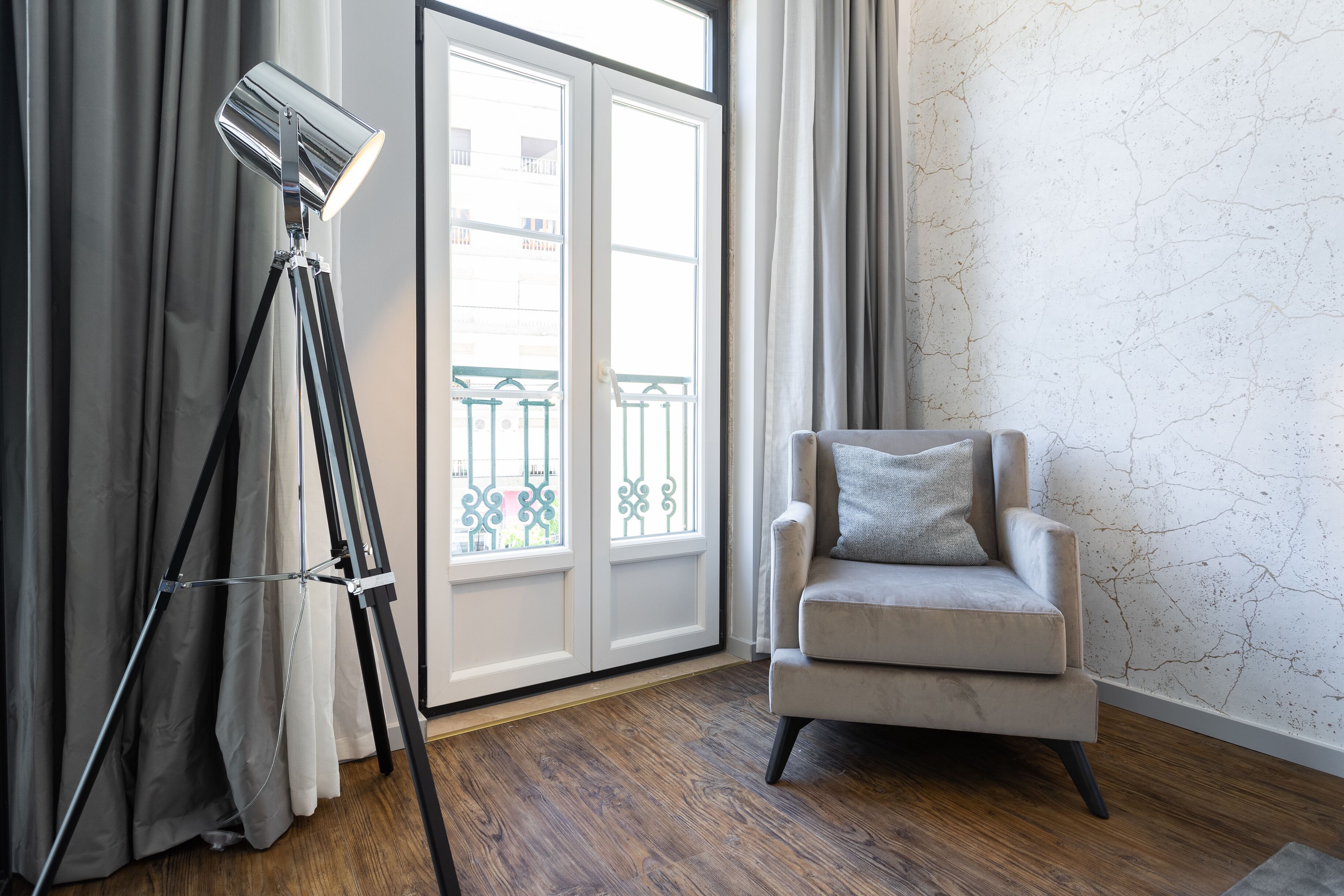 Une pièce comprenant un lampadaire sur trépied, un fauteuil beige avec un coussin, une grande fenêtre avec des rideaux et un plancher en bois.