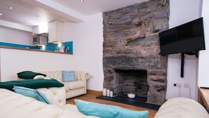 A living room with a stone fireplace, a flat-screen TV mounted on the wall, and a sofa with cushions. - The Captain's Bank (Porthmadog)