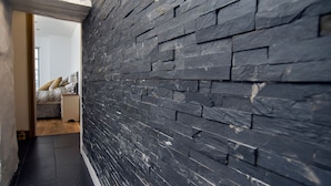 A hallway with a dark stone wall, a wooden floor, and a bed with a nightstand in the background. - The Captain's Bank (Porthmadog)