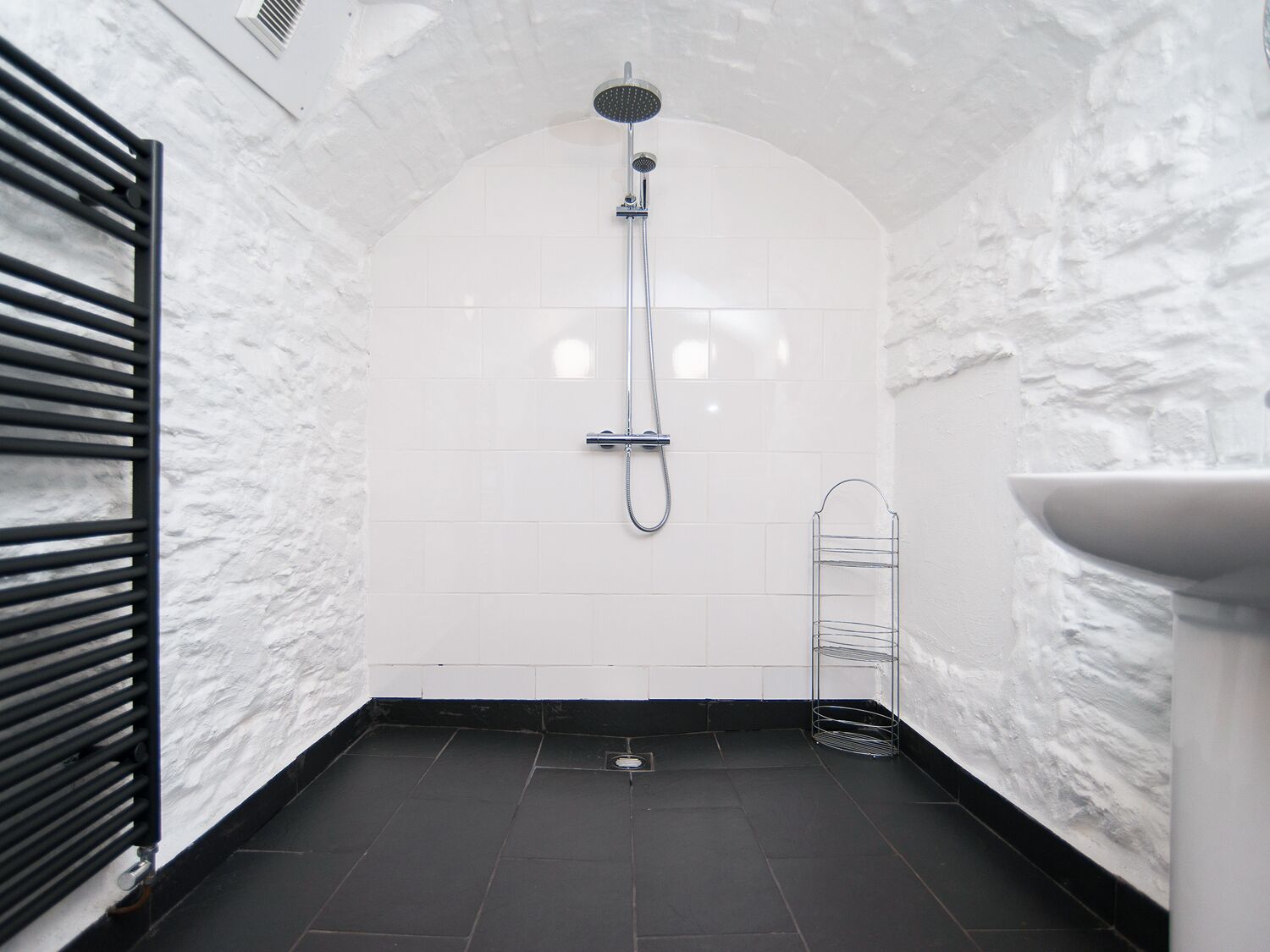 A small, white-tiled bathroom with a shower, a black radiator, and a marble countertop.