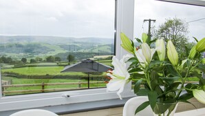 A view from a window with a table and chairs, a vase with white flowers, and a landscape of green fields and hills. - Caesamson (Caersws)