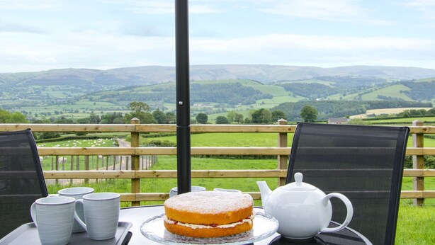 A table with a cake, teapot, and cups, overlooking a scenic countryside view. - Caesamson (Caersws)