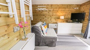 A modern room with a wooden wall, a bed with a colorful blanket, a white dresser, a flat-screen TV, and a small table with flowers. - Cilan Lodge (Corwen)
