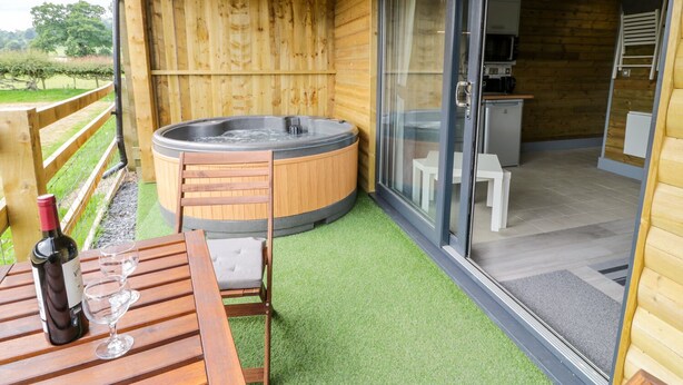 A wooden deck with a hot tub, a table with a bottle of wine and a glass, and a sliding glass door leading to a kitchen area. - Cilan Lodge (Corwen)