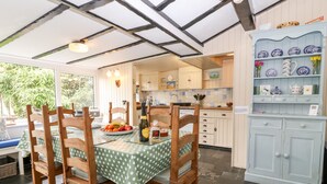 Uma sala de jantar com uma mesa posta para quatro pessoas, rodeada por cadeiras de madeira. Uma cozinha com armários brancos e um armário azul com objetos decorativos é visível ao fundo.