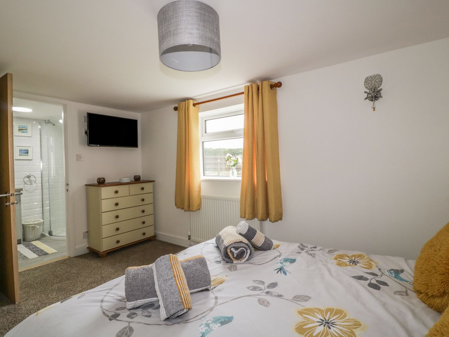 A bedroom with a bed, a dresser, a television, a window with curtains, and a wall-mounted light fixture.