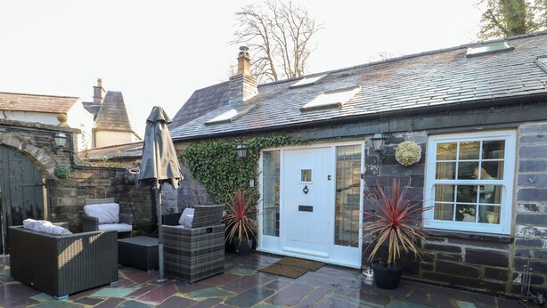A stone patio with outdoor seating, a white door, and a window with a view of the interior. - Bryn Derwen Coach House (Bangor)