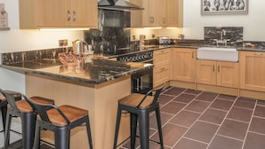 A modern kitchen with wooden cabinets, a black stove, and a marble countertop. There are two bar stools and a sink. - Bryn Derwen Coach House (Bangor)