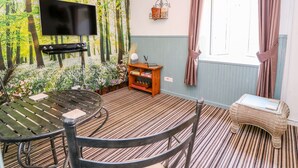A room with a forest-themed wall mural, a flat-screen TV, a round table with chairs, a small wooden side table, and a wicker bench. - River Cottage (Port Talbot)