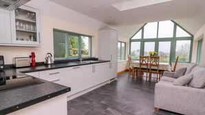 Una cocina moderna con armarios blancos, encimera negra y estufa empotrada. Hay un comedor con un sofá gris y una mesa con sillas. Amplias ventanas ofrecen luz natural y vistas al exterior.