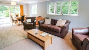 A spacious living room with a brown leather sofa, a wooden coffee table, and a dining area with a wooden table and chairs. Large windows provide natural light and a view of greenery outside. - Tanglewood (Colwyn Bay)