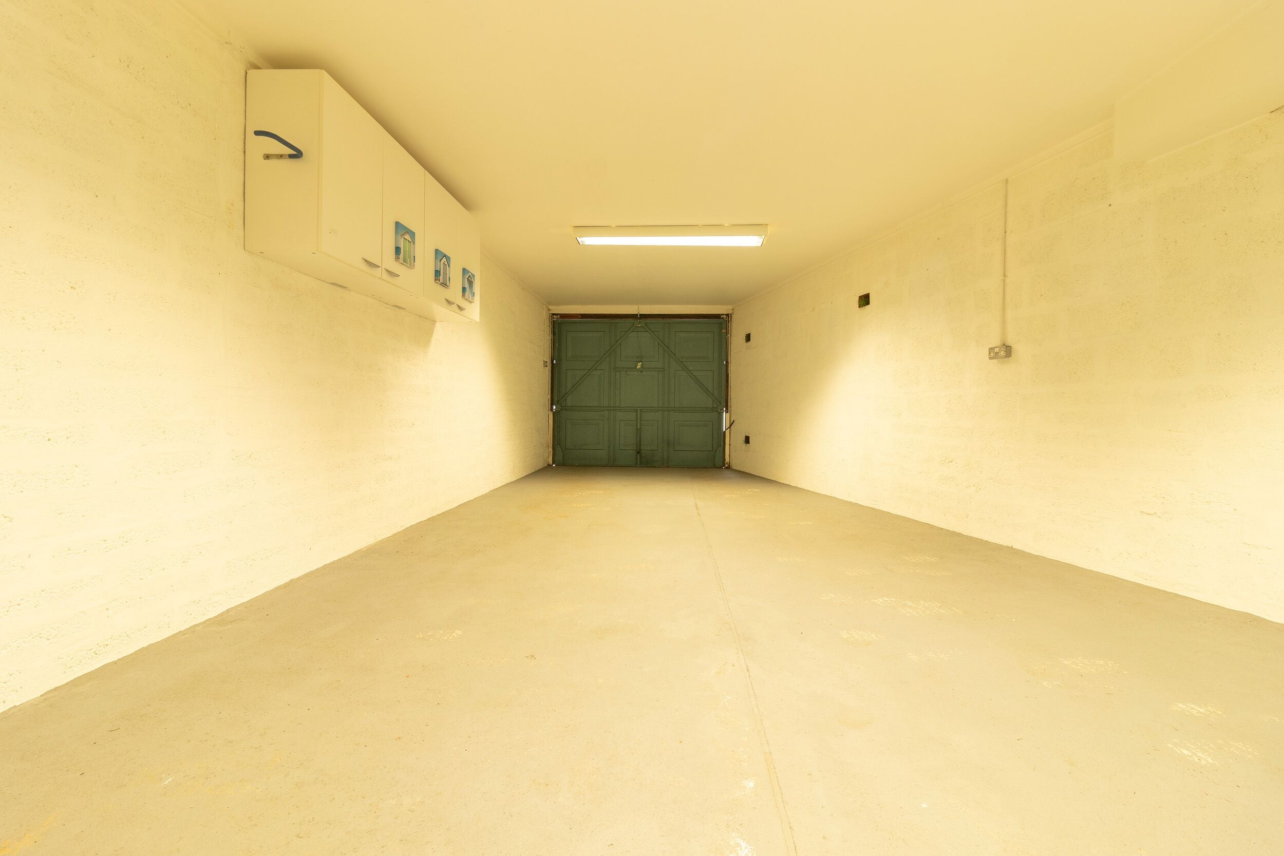 A narrow hallway with a green door at the end, a light fixture on the ceiling, and a wall-mounted cabinet with a blue handle.
