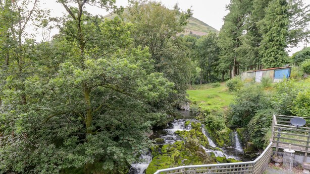 A serene outdoor area with a small waterfall, surrounded by lush greenery and a wooden deck with a table and chairs. - Dyfi Cottage (Machynlleth)